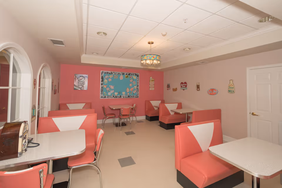 A retro-style dining room with pink and white booth seating and tables. The walls are painted pink and decorated with vintage-style signs and a bulletin board. There are also some metal chairs and tables in the background, and a ceiling light fixture with a colorful shade.