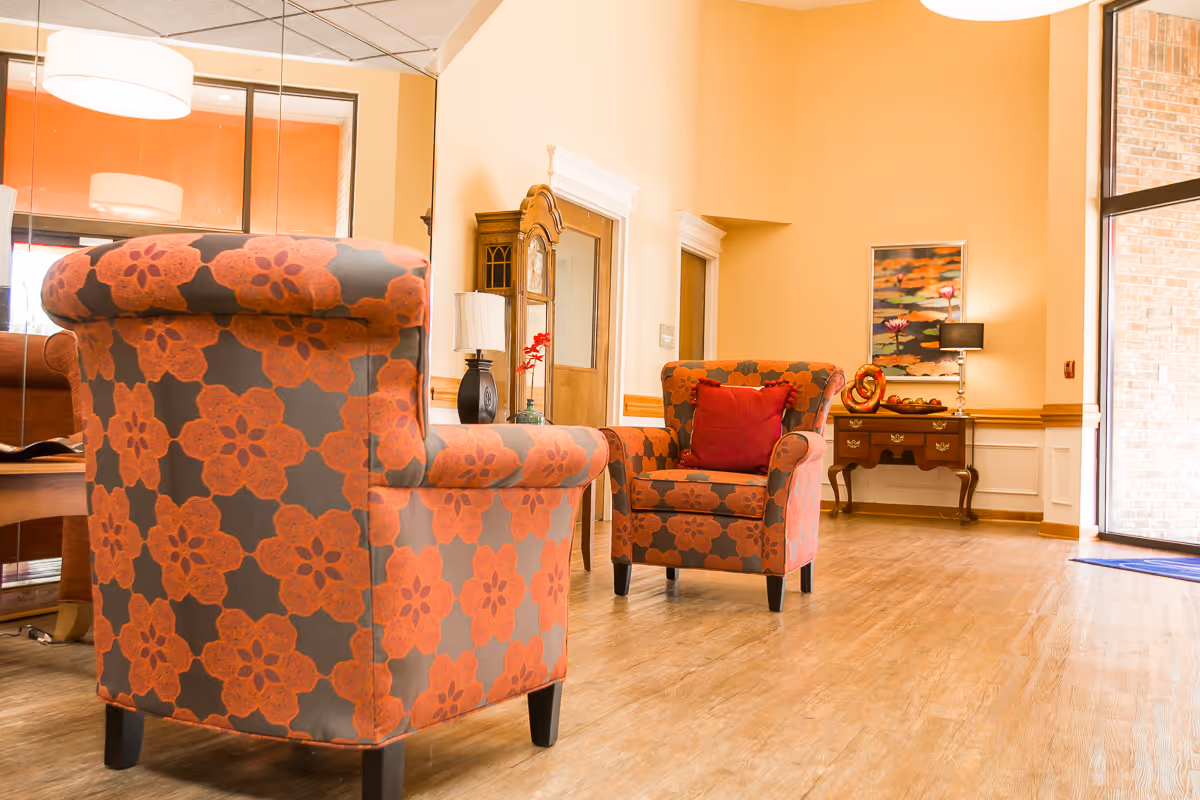 A bright and welcoming interior seating area with two orange and gray floral patterned armchairs, one with a red cushion. The room has light wooden flooring, beige walls, a wooden side table with decorative items and a lamp, a large mirror, and a glass door letting in natural light.