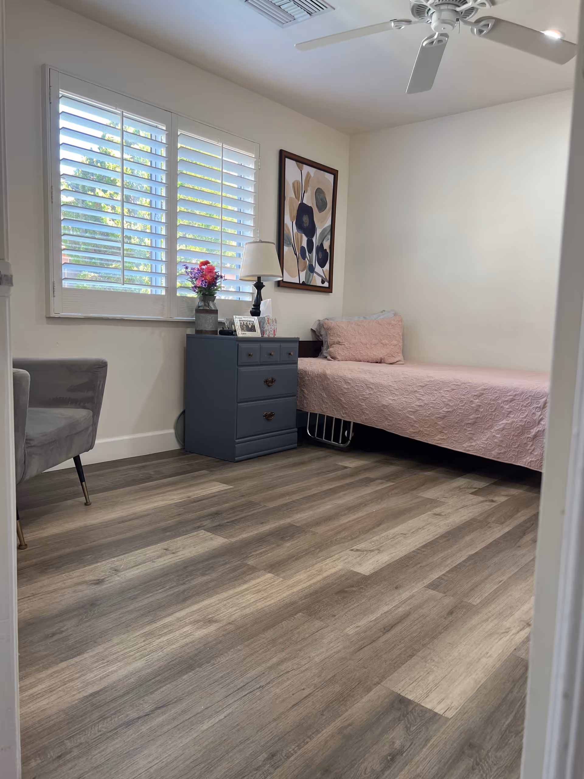 Small bedroom with a single bed covered in a pink quilt, a blue bedside dresser with a lamp and flowers, a gray chair, and a window with white shutters.
