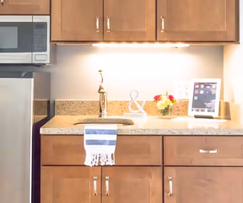 A kitchen countertop with a built-in sink and faucet, wooden cabinets above and below, a microwave oven mounted above a stainless steel refrigerator, a striped towel hanging from the cabinet handle, a small flower arrangement, and a tablet on a stand on the counter.