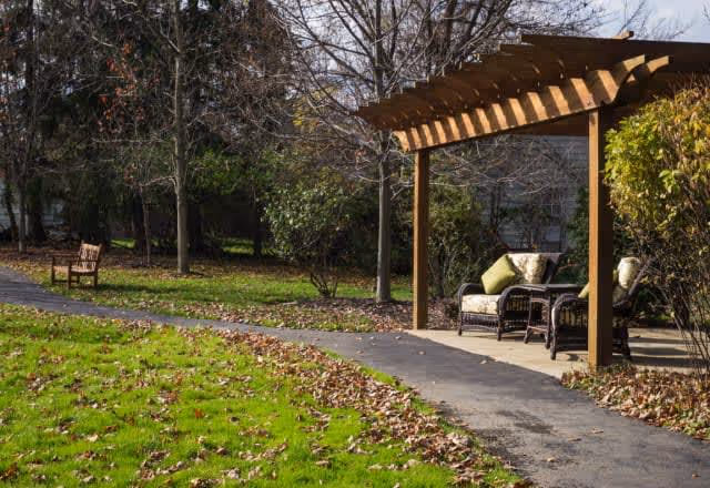 Outdoor garden area with a paved walkway, a wooden pergola with cushioned wicker chairs and a small table underneath, surrounded by trees and bushes with fallen leaves on the grass.