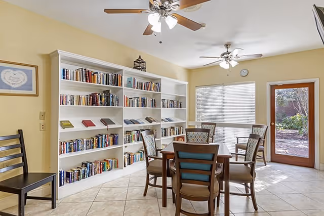 Bright communal reading and activity room with bookshelves, a table and chairs, ceiling fans, and a door to an outdoor area.