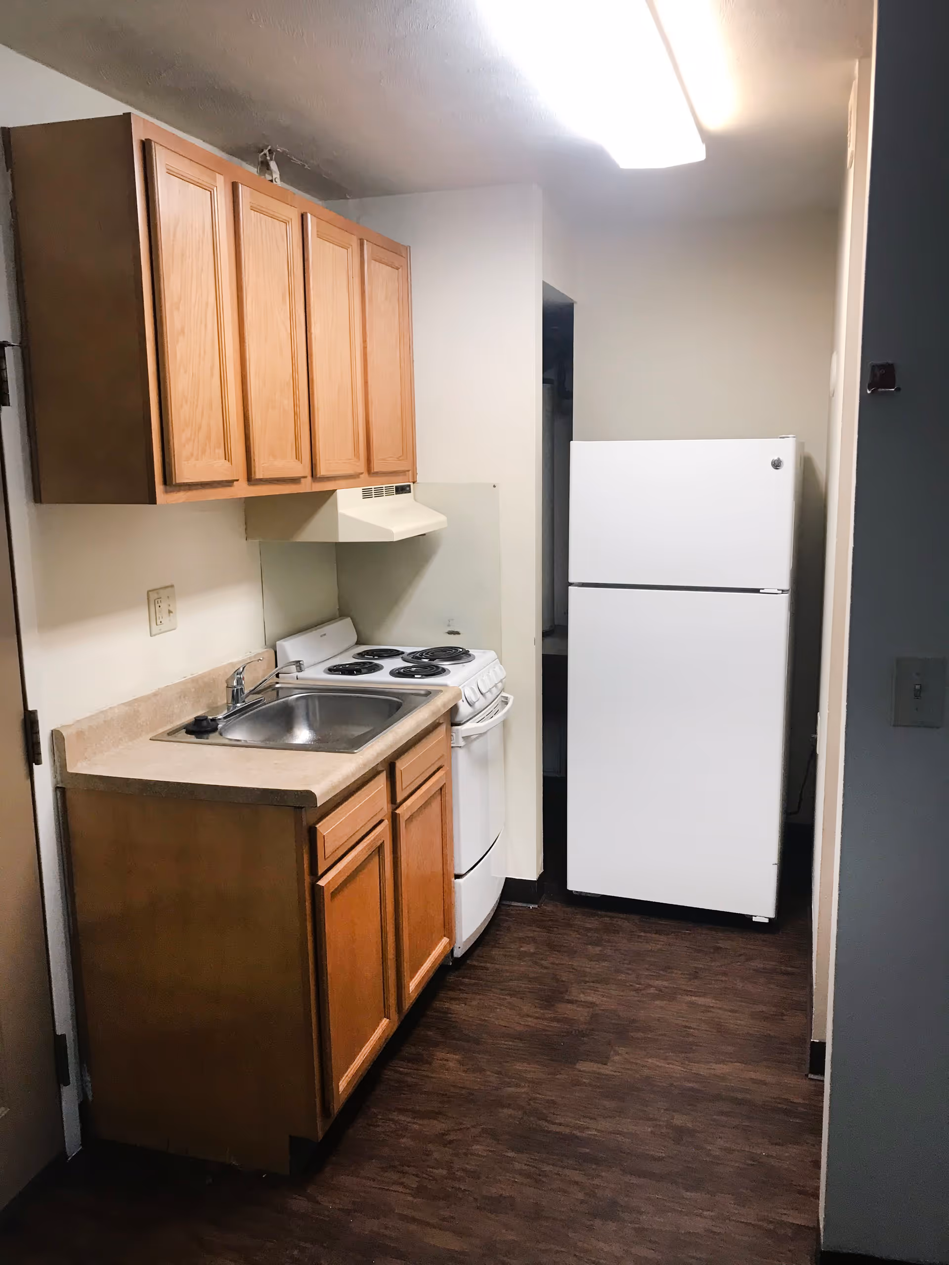 Small kitchen area with wooden cabinets, a stainless steel sink, a white electric stove with four burners, a white refrigerator, and wood-patterned flooring under bright ceiling light.