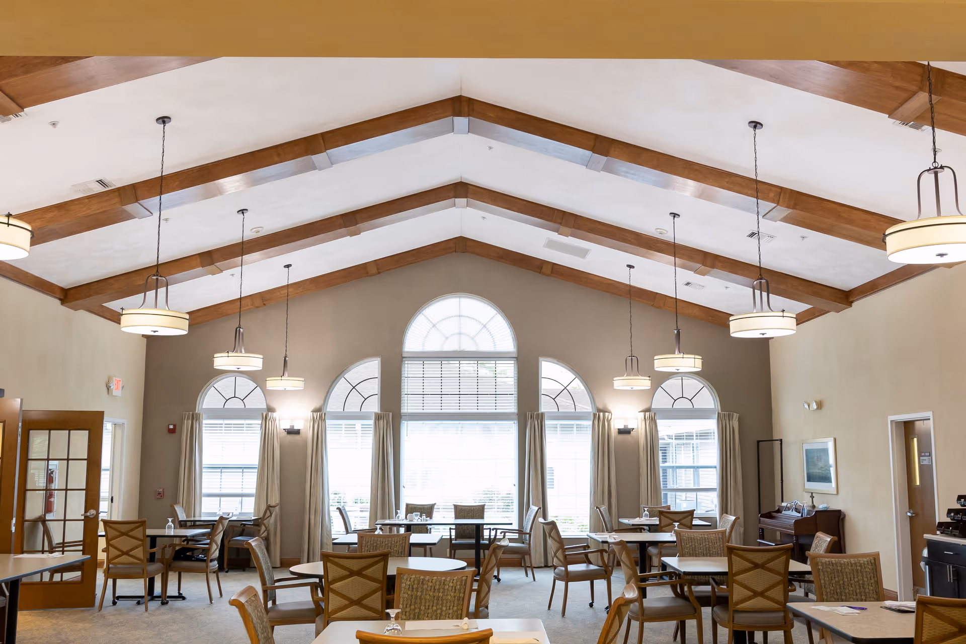 A bright dining room with multiple round tables and chairs arranged neatly. The room features large arched windows with curtains, wooden ceiling beams, and several hanging pendant lights. The walls are painted a warm beige color, and there is a door and some furniture visible on the right side.