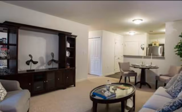 A cozy living room area with beige carpet, a dark wooden entertainment center, a glass-top coffee table with books and decorative items, two armchairs, and a sofa with patterned cushions. In the background, there is a small dining area with a round table set for two and a kitchen with white cabinets and stainless steel appliances.