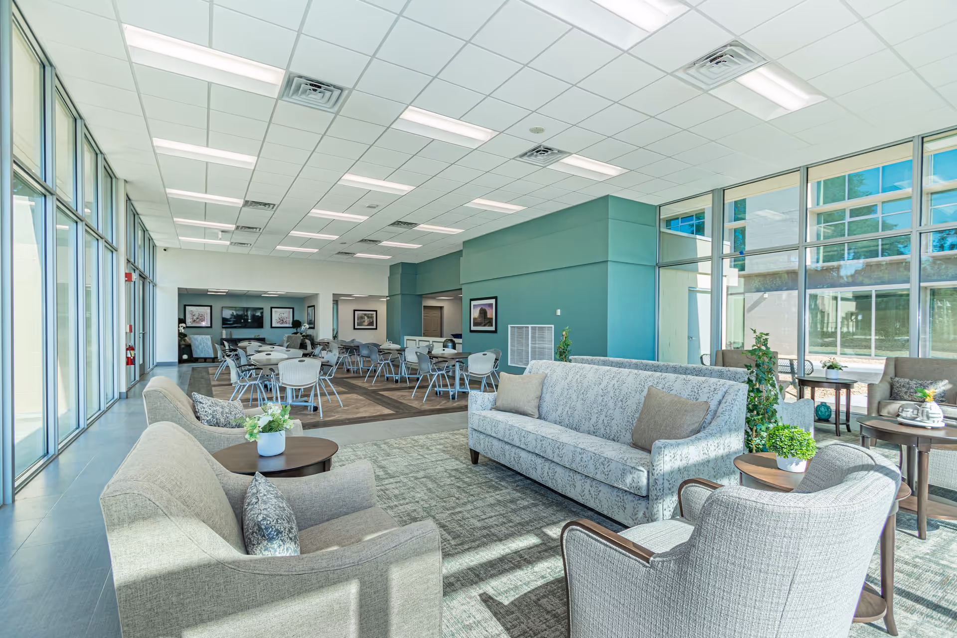 A bright and spacious common area in a senior living facility with large windows letting in natural light. The room features comfortable seating including sofas and armchairs arranged around small tables with plants and flowers. In the background, there are multiple round tables with chairs, and framed artwork on the walls. The ceiling has recessed lighting and air vents.