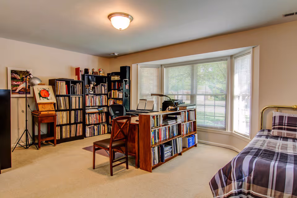 A cozy room with a large bay window letting in natural light. The room features a bed with a plaid bedspread on the right, a wooden desk with bookshelves filled with books and records, a chair, and a tall bookshelf against the wall. There is also a floor lamp and framed artwork on the left side.