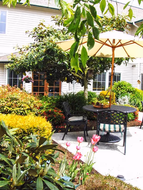 Outdoor patio area at a senior living facility with a round metal table and two chairs, one with a striped cushion. A large beige umbrella shades the table. Surrounding the patio are various green plants, bushes, and pink tulips, with a white building featuring multiple windows in the background.