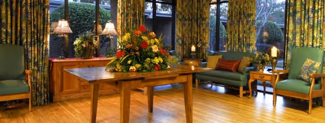 A cozy lounge with a wooden table holding a large floral arrangement, green upholstered chairs and sofas, lamps, and patterned floor-to-ceiling curtains.