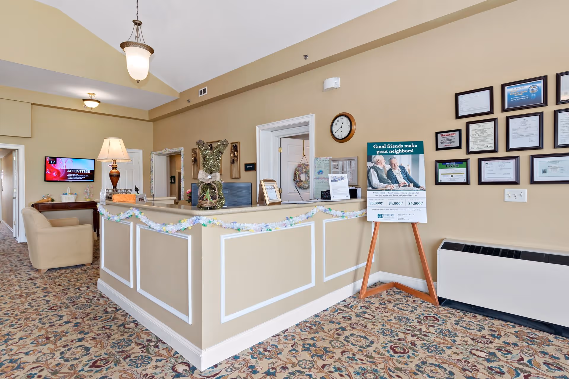 Reception area of Bentley Assisted Living at Branchville with a beige front desk decorated with a garland, a lamp, and a decorative bunny figure. Behind the desk are doors, a clock, and a wall with multiple framed certificates and awards. A sign on an easel promotes referral bonuses. There is a patterned carpet and a beige armchair near a wall-mounted TV displaying activities.