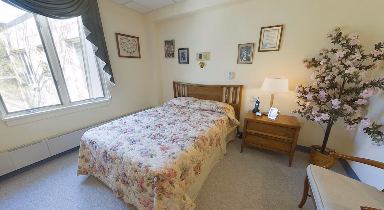 A cozy bedroom with a double bed covered in a floral bedspread. There is a wooden nightstand with a lamp, a phone, and a framed photo. The walls have several framed pictures and certificates. A large window with drapes lets in natural light. A potted flowering plant and a cushioned chair are also visible in the room.