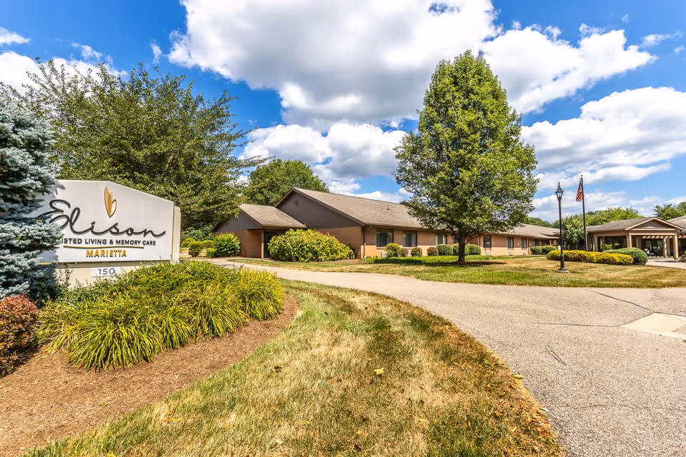 Exterior view of Elison Assisted Living & Memory Care of Marietta facility on a sunny day with a partly cloudy sky. The building is single-story with a brown roof and beige walls, surrounded by green trees and bushes. A paved driveway curves in front of the building, and there is a sign with the facility's name near the entrance. Two flagpoles with an American flag and another flag are visible near the building entrance.