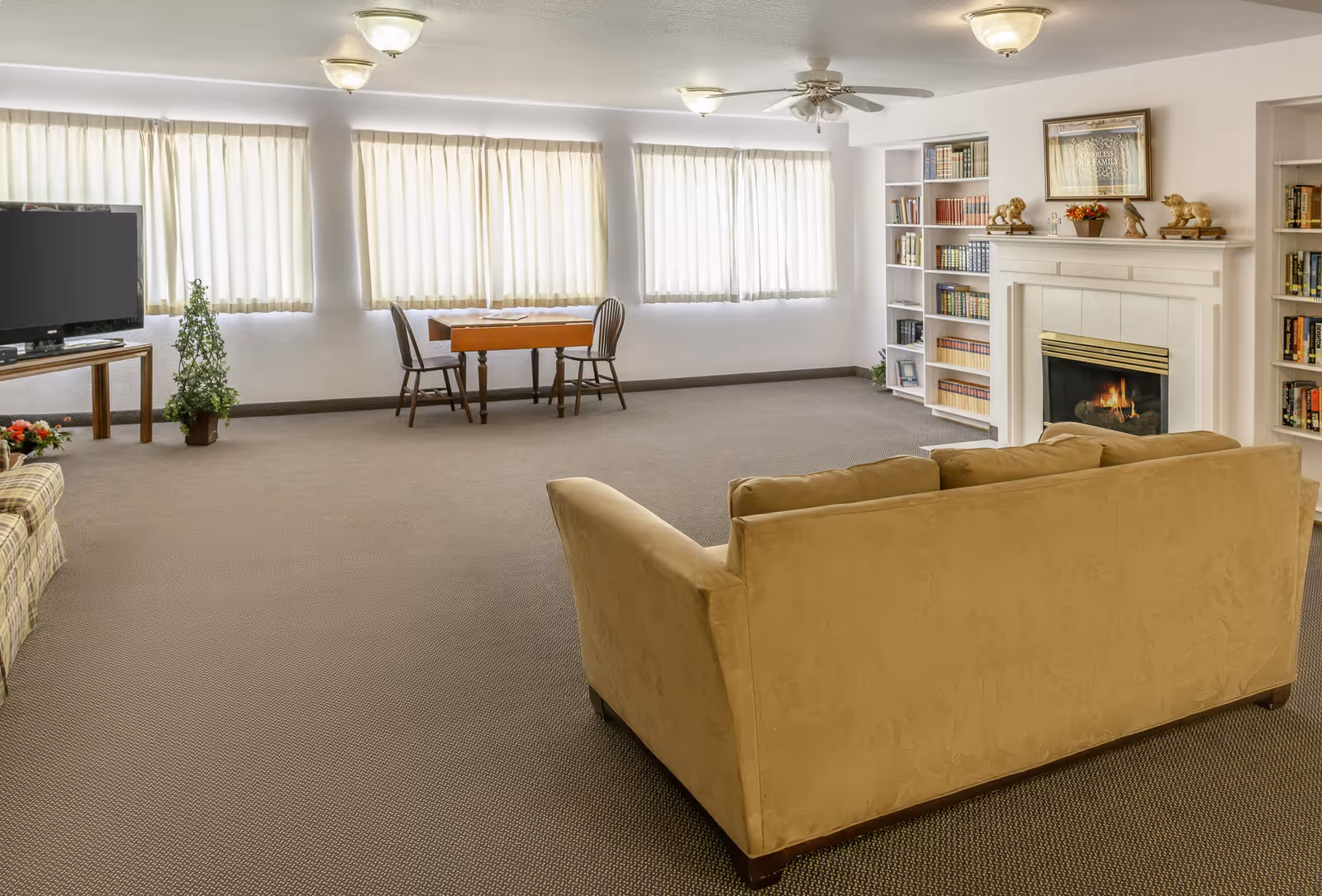 Spacious common room with a sofa facing a fireplace and bookshelves, a TV, and a small table by sunlit windows.