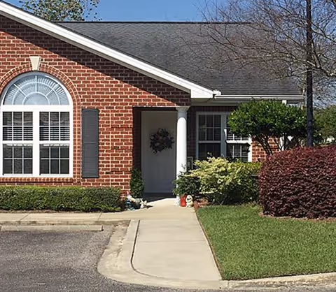 Exterior view of a brick building with a large arched window and a white door decorated with a wreath. There is a concrete walkway leading to the entrance, surrounded by green bushes and a well-maintained lawn under a clear sky.
