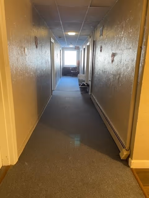 A long indoor hallway with beige walls and a gray carpeted floor. Several doors line both sides of the hallway. At the end of the hallway, there is a window letting in natural light and a small table with a chair. The ceiling has recessed lighting and a drop ceiling design.