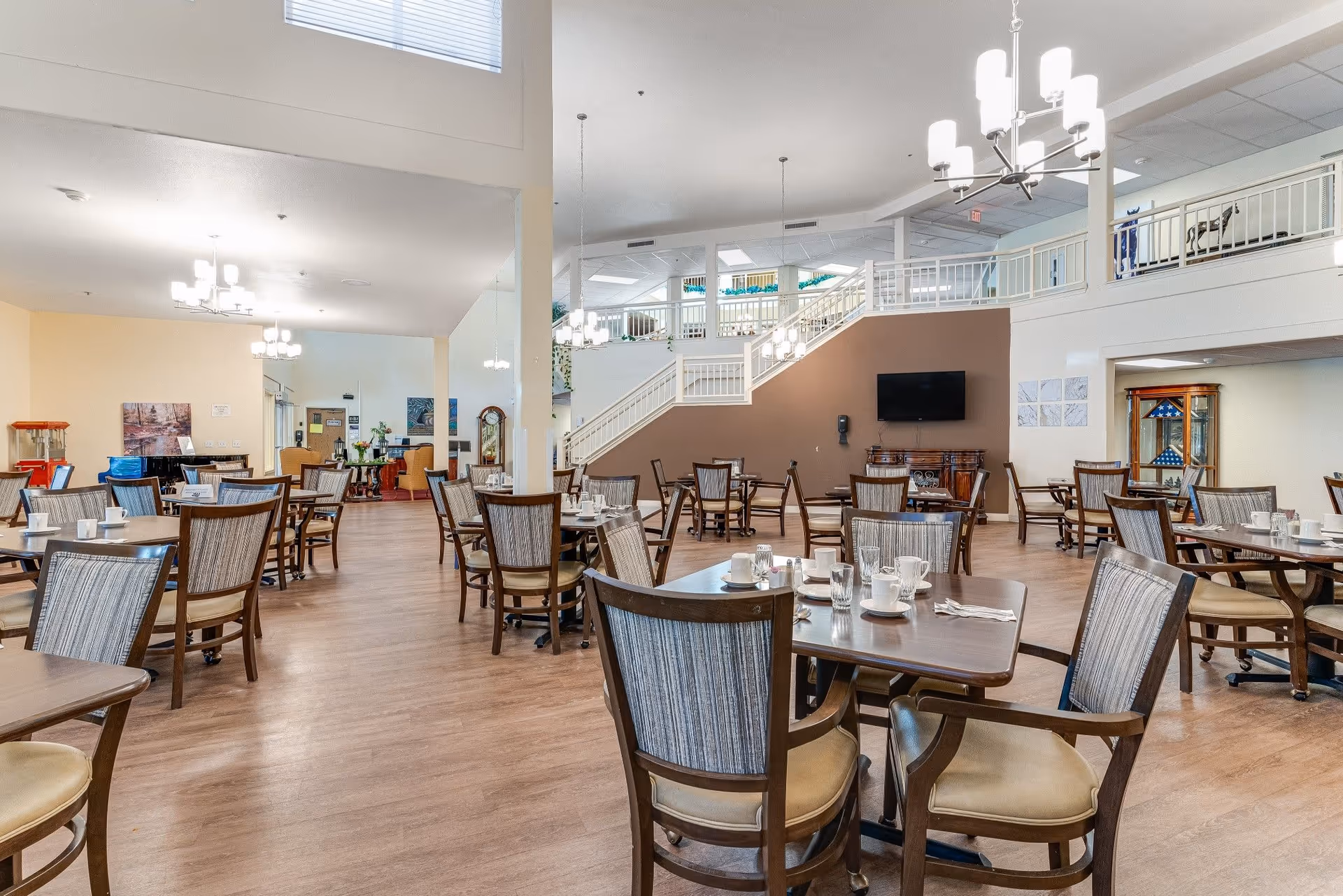 A spacious dining room in a senior living facility with multiple wooden tables and chairs arranged neatly. Each table is set with cups, glasses, and napkins. The room has wooden flooring, modern chandeliers, a staircase leading to an upper level, and a television mounted on a brown accent wall.