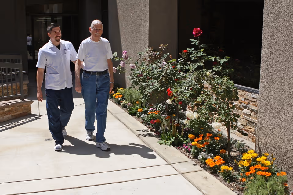 A caregiver in a white uniform walks alongside an elderly man wearing a white t-shirt and jeans on a sunny outdoor pathway next to a flower bed with colorful flowers and green plants.