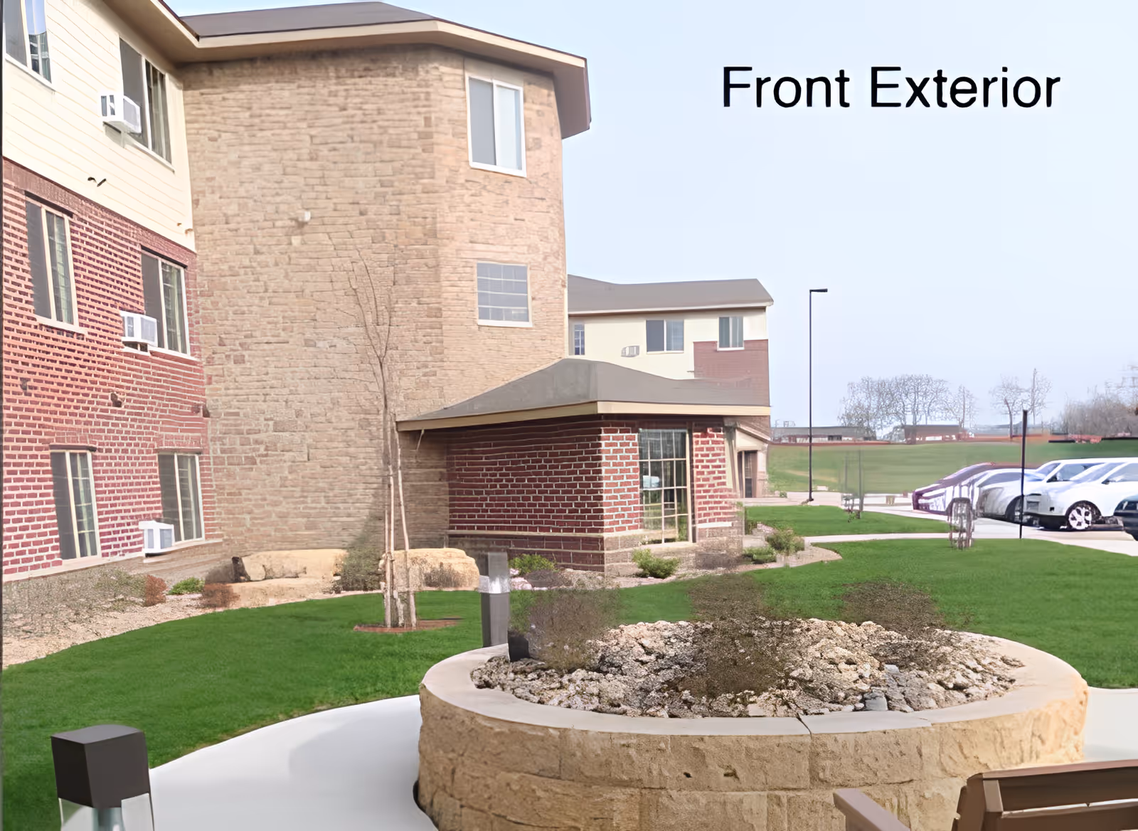 Front exterior of a multi-story building with brick and stone facade, landscaped courtyard and parked cars.
