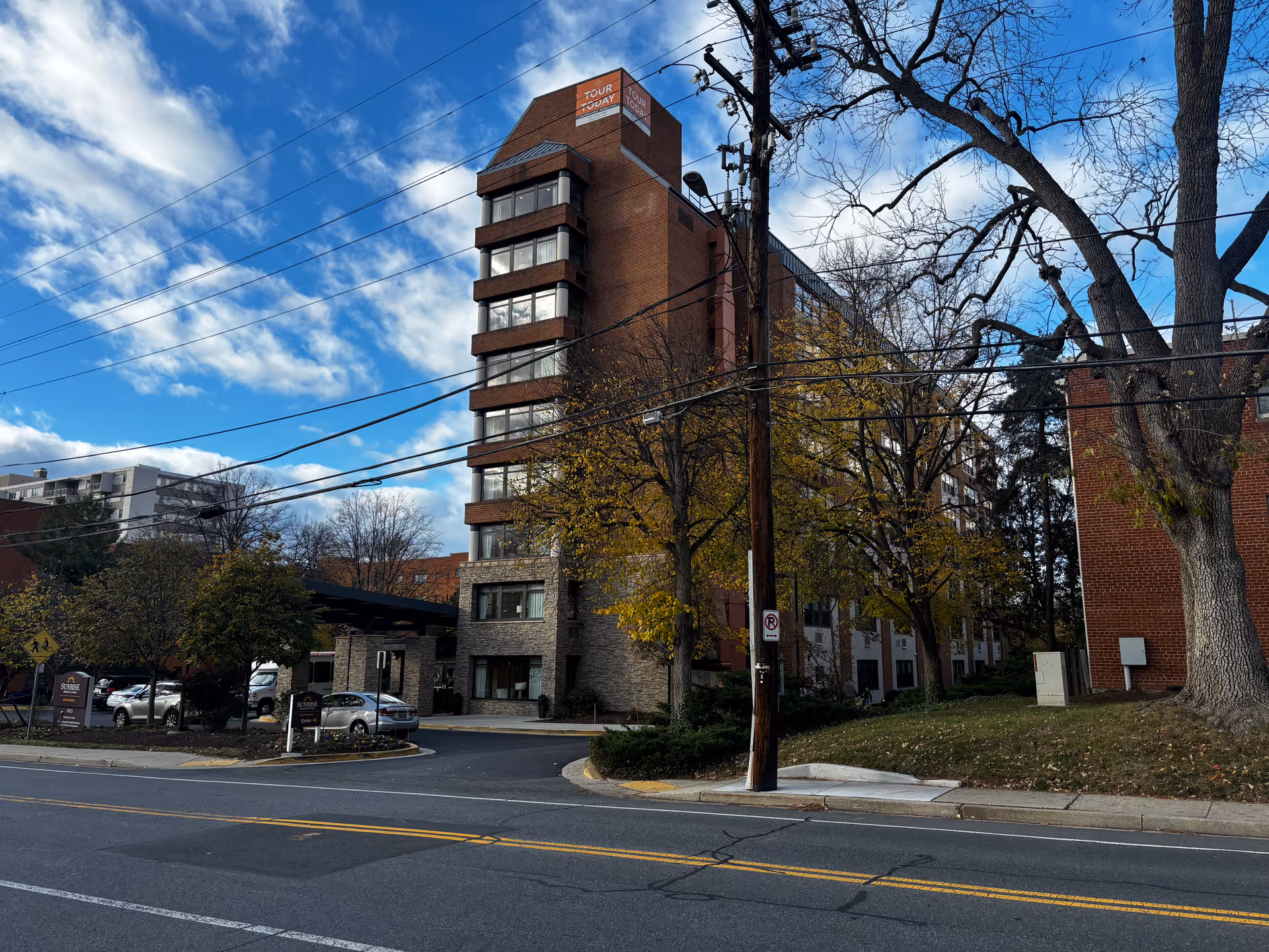 Exterior view of a multi-story brick building with a covered entrance and signs for Sunrise of Bethesda. The scene includes a street in the foreground, trees with autumn leaves, and a partly cloudy blue sky.