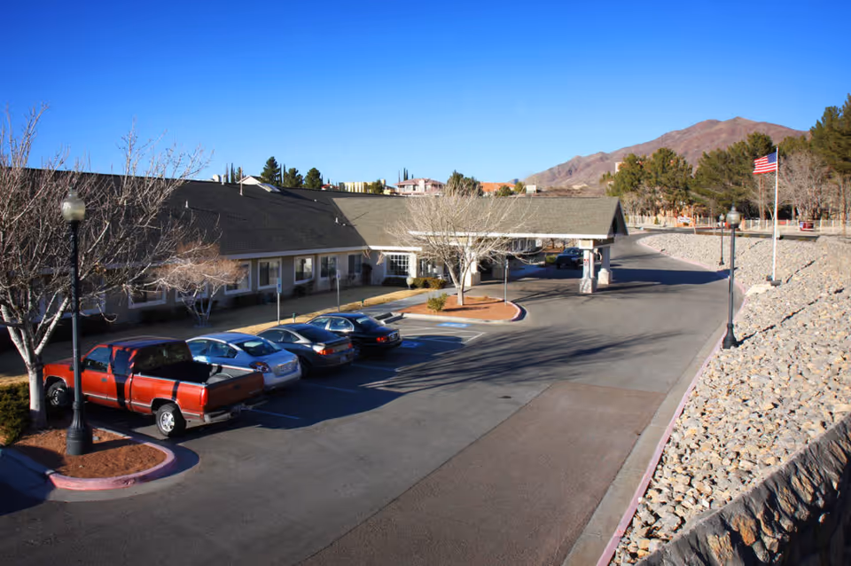 Exterior view of SunRidge at Desert Springs facility showing a parking lot with several parked cars, a covered entrance, leafless trees, lamp posts, an American flag on a flagpole, and mountains in the background under a clear blue sky.