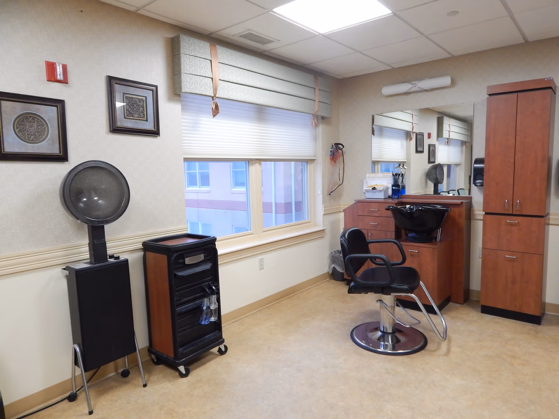 Interior view of a hair salon area in a senior living facility with a black salon chair, a hair washing sink, wooden cabinets, a rolling cart with hair products, a hair dryer, two framed decorative pictures on the wall, and a window with a valance and blinds.