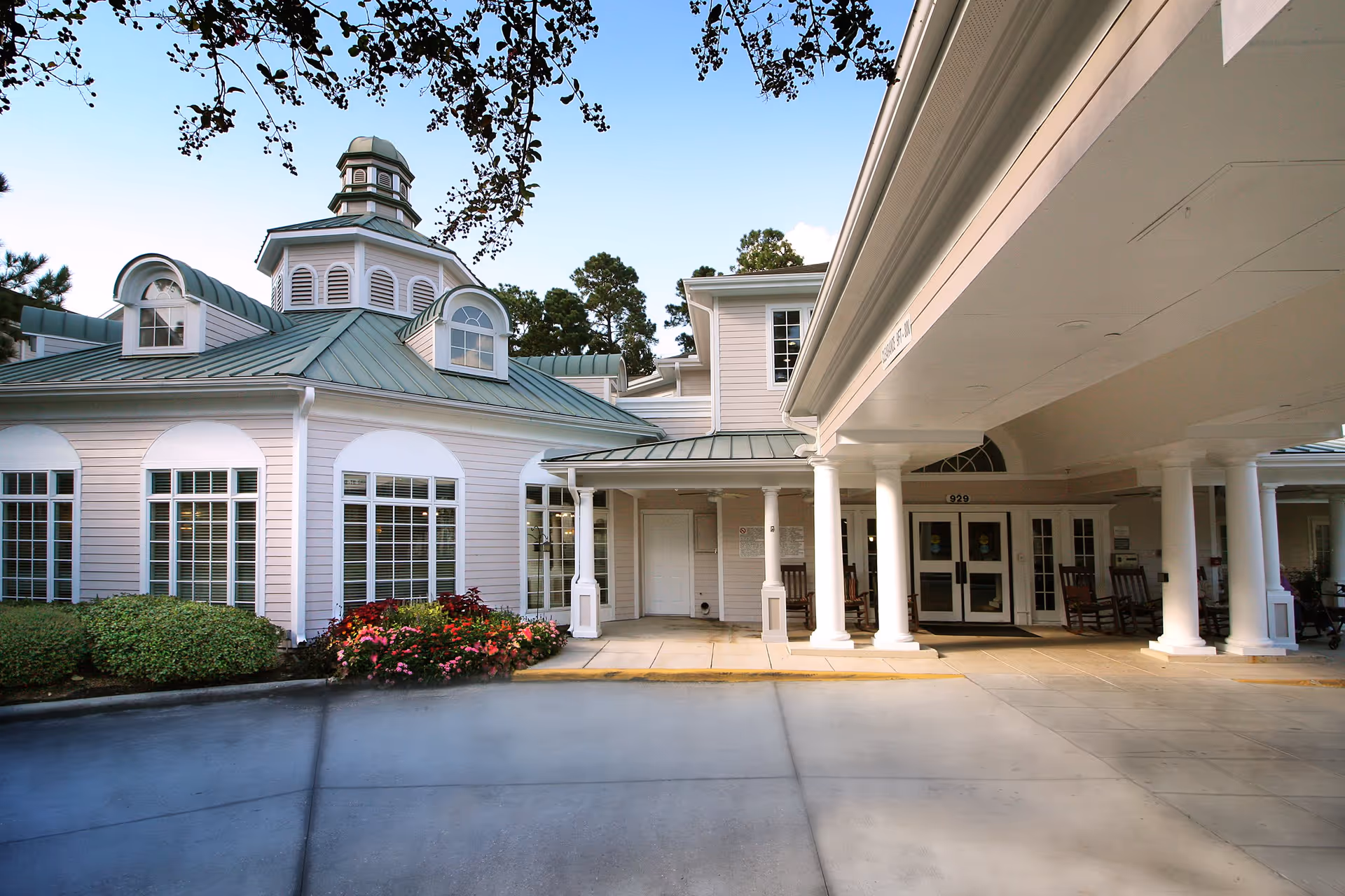 Exterior view of King's Preserve at Kingwood senior living facility showing a covered entrance with white columns, rocking chairs, and a building with multiple windows and a green metal roof under a clear blue sky.
