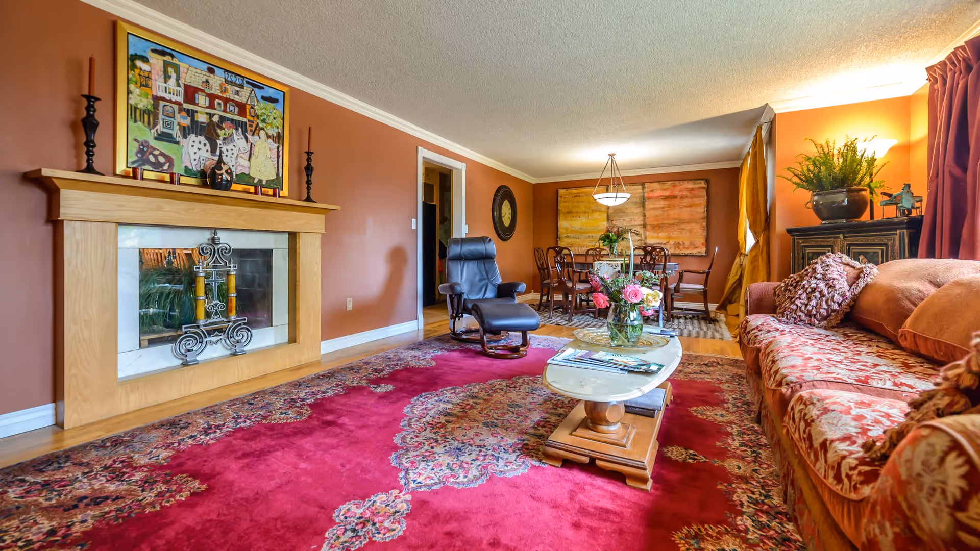 A cozy living room with a red patterned carpet, a wooden fireplace with decorative candles, a floral-patterned sofa with cushions, a glass coffee table with flowers and magazines, a black recliner chair, and a dining area with a wooden table and chairs in the background. The walls are painted a warm brown color and decorated with artwork and a clock.