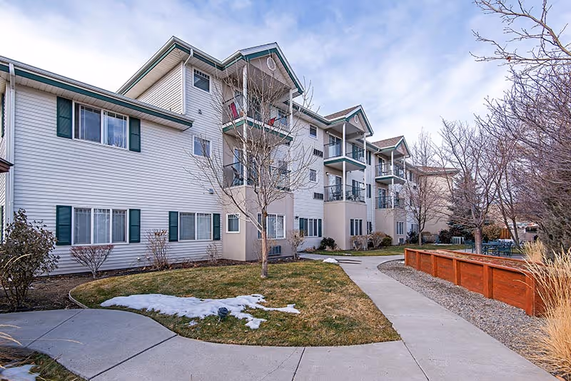Exterior view of a multi-story senior living facility building with white siding and green shutters. There are balconies on the upper floors and a paved walkway leading through a landscaped area with grass, leafless trees, and some patches of snow.