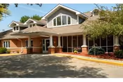 Exterior view of a single-story assisted living facility with a covered entrance, large windows, and a well-maintained driveway surrounded by greenery and trees.
