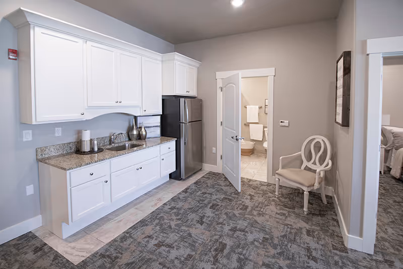A small assisted-living kitchenette with white cabinets, granite countertop, a stainless refrigerator, and an open door leading to a bathroom and bedroom.