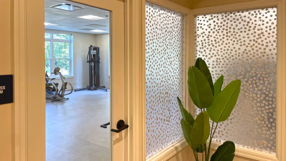 View through an open door into a fitness room with exercise equipment including a stationary bike and a multi-function gym machine. The door has a black handle and a sign partially visible on the left side. To the right of the door, there is a corner window with frosted glass featuring a floral pattern and a green leafy plant in front of it.