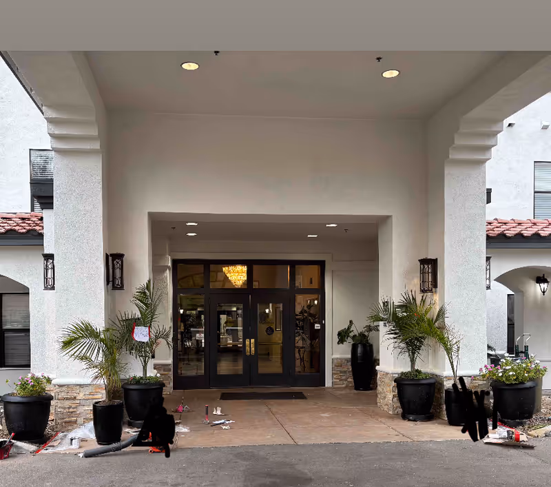 Entrance of a senior living facility with double glass doors under a covered driveway. There are potted plants on either side of the entrance and a chandelier visible inside through the glass doors. The building has white walls and a tiled roof.