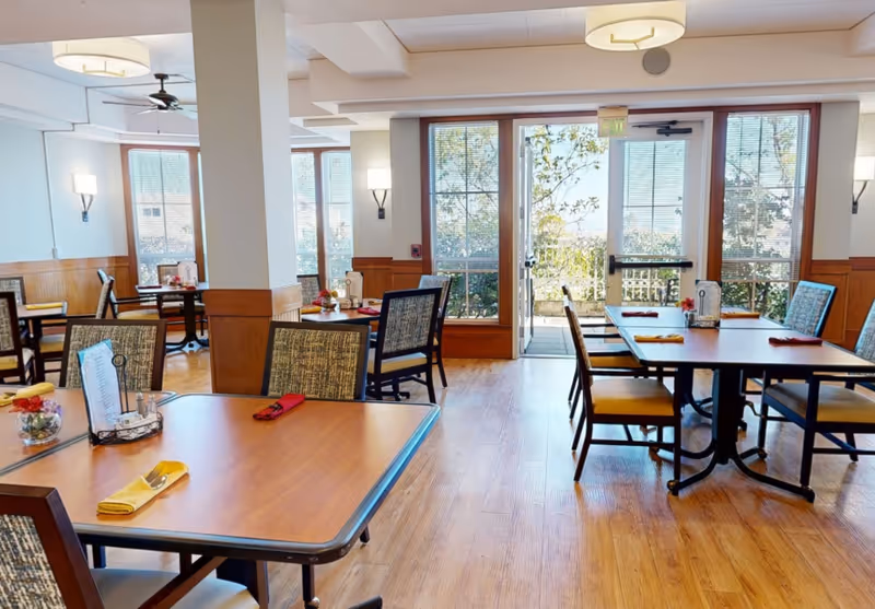 A bright dining room with wooden floors and several tables set with yellow and red napkins. Chairs with patterned upholstery surround the tables. Large windows and a glass door let in natural light and provide a view of greenery outside.