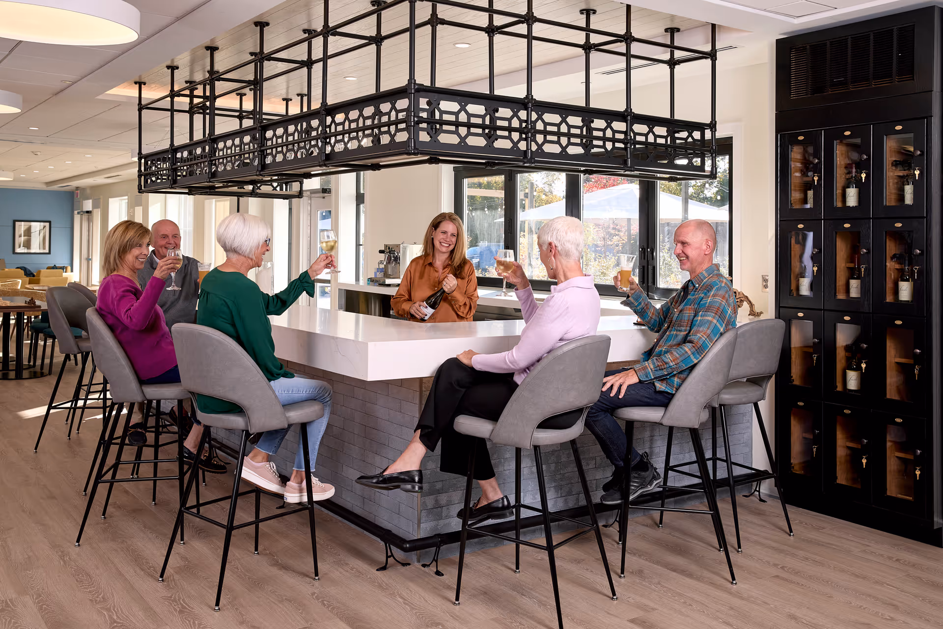 A group of five seniors sitting on bar stools around a modern indoor bar counter, raising their glasses in a toast with a bartender smiling behind the counter. The room has large windows, wooden flooring, and a wine storage cabinet on the right.