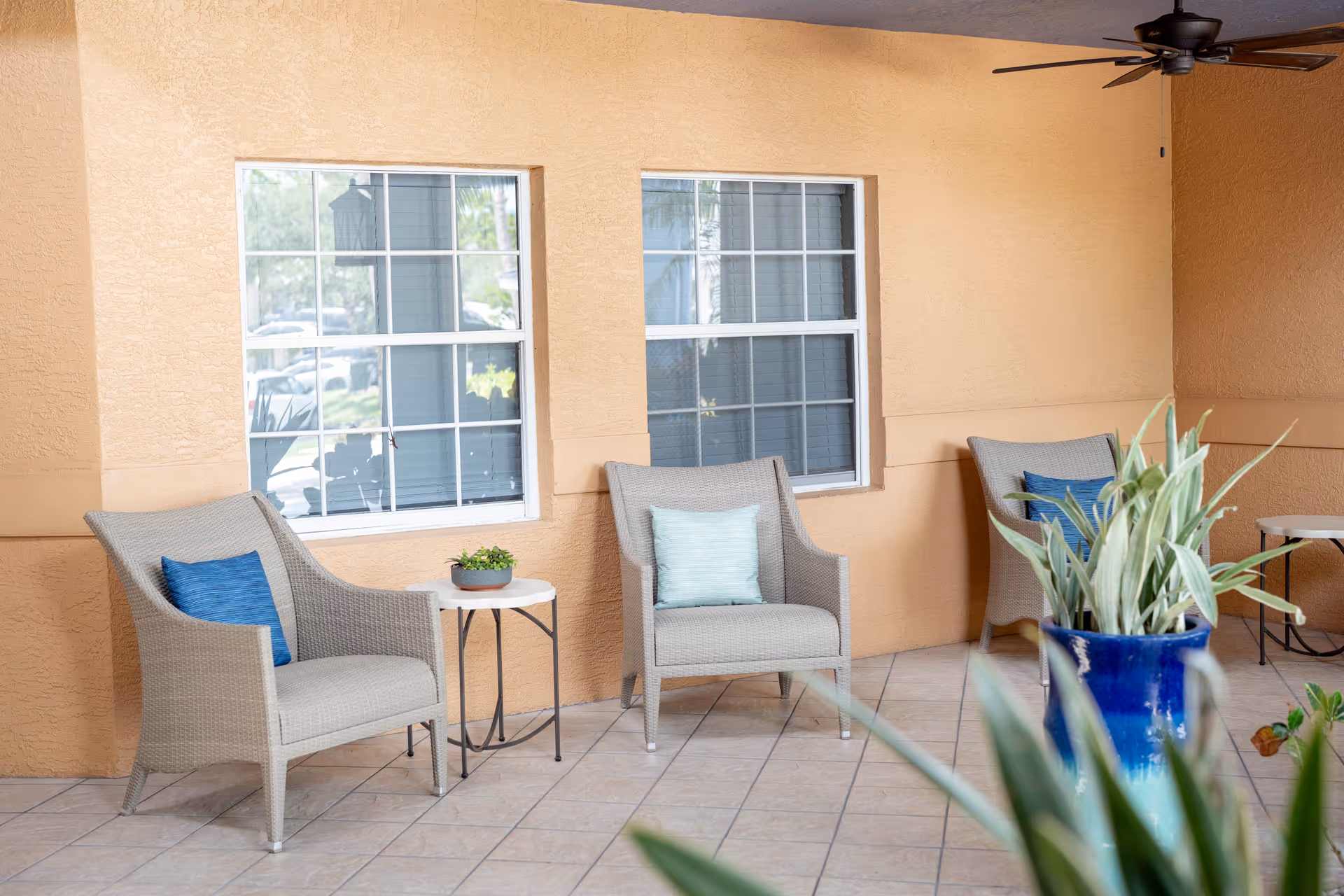 A covered outdoor patio area with three beige wicker chairs, each with a blue or light green cushion. There are two small round white tables, one with a small potted plant. The walls are painted a warm beige color, and there are two windows with white frames. A ceiling fan is mounted on the ceiling, and a large blue ceramic pot with green plants is in the foreground.