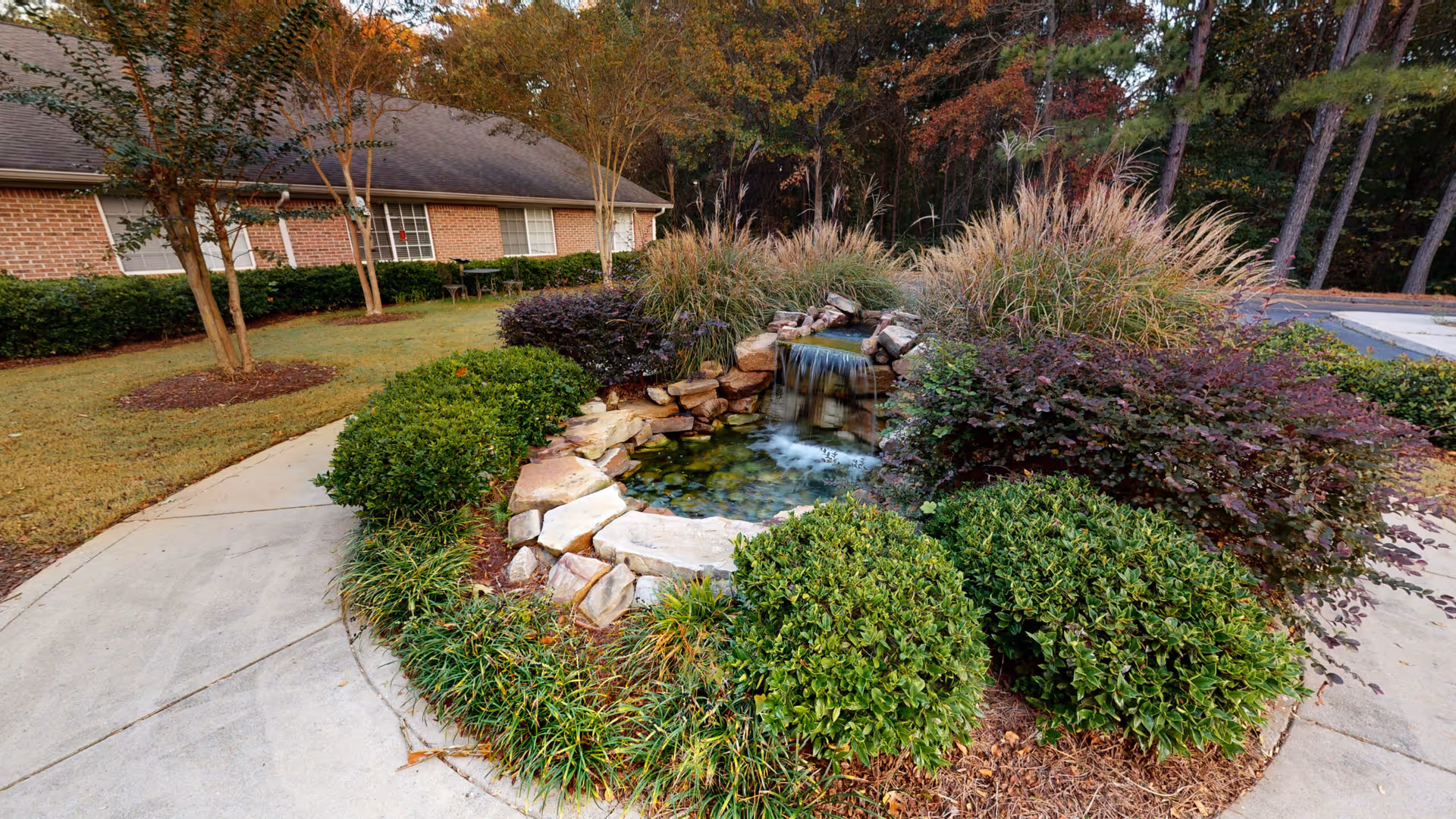 Landscaped outdoor courtyard with a small rock waterfall pond, trimmed shrubs, and a curved concrete walkway in front of a brick building.