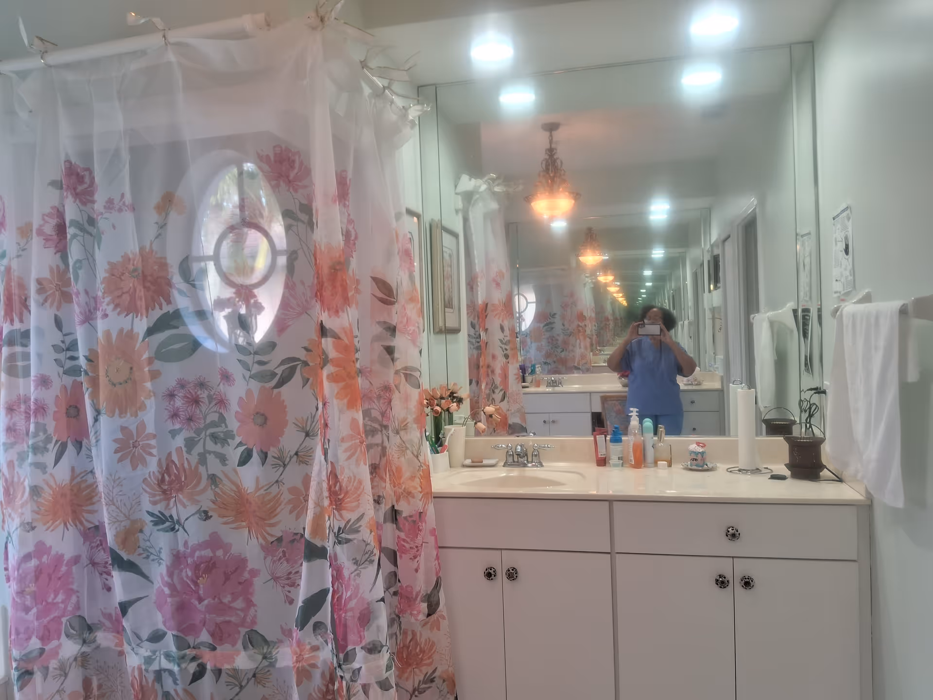 A bathroom with a floral shower curtain featuring pink and orange flowers. There is a white countertop with a sink, various toiletries, and a paper towel roll. A large mirror reflects the room and a person taking the photo. The bathroom has white walls, towel racks with white towels, and a decorative light fixture on the ceiling.