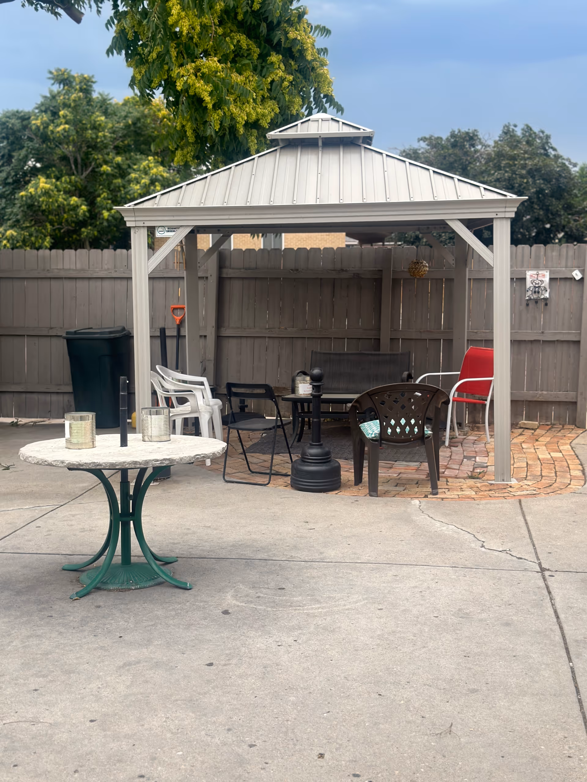 Outdoor patio area with a small gazebo structure containing several chairs and a table. The area is enclosed by a wooden fence, and there is a round table with a green base and two tin cans on it in the foreground. Trees and a partly cloudy sky are visible in the background.