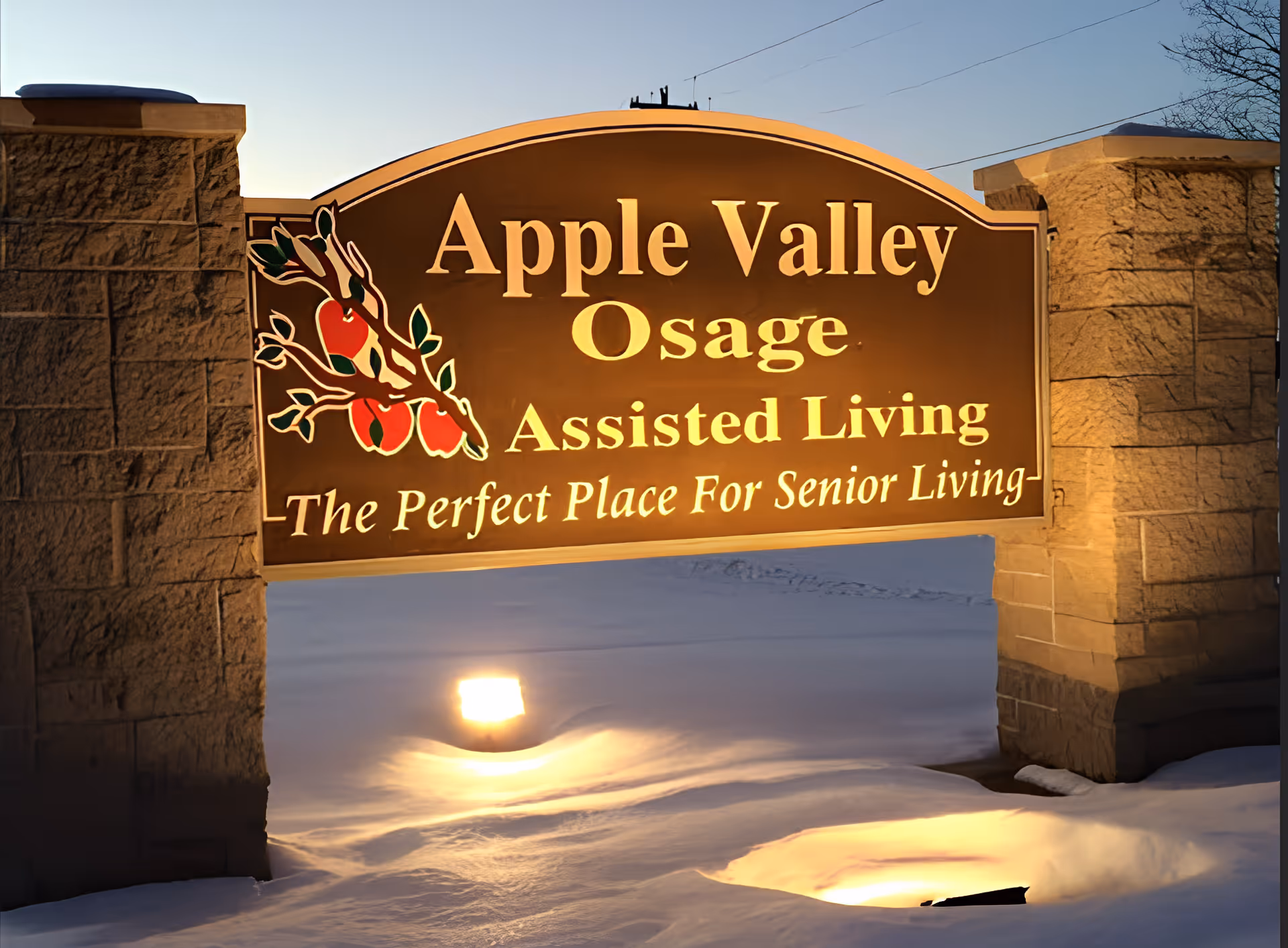 Illuminated sign for Apple Valley Osage Assisted Living with a decorative branch of apples on the left side, set between two stone pillars with snow on the ground at dusk.
