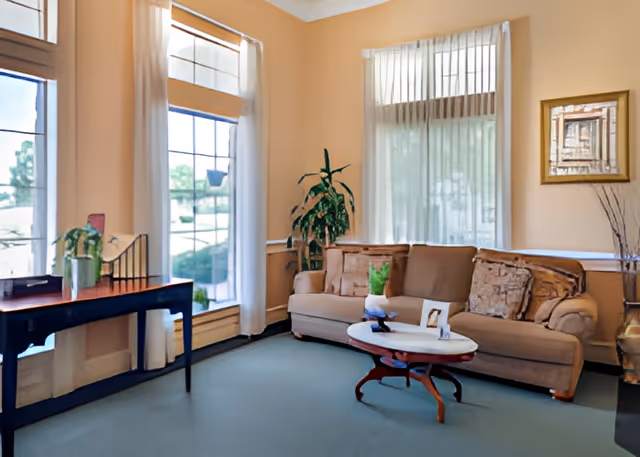 A cozy living room with a beige sofa adorned with patterned cushions, a round wooden coffee table with decorative items, a tall green plant in the corner, large windows with white curtains allowing natural light to fill the room, a wooden table with a plant and other items near the windows, and a framed artwork on the wall.