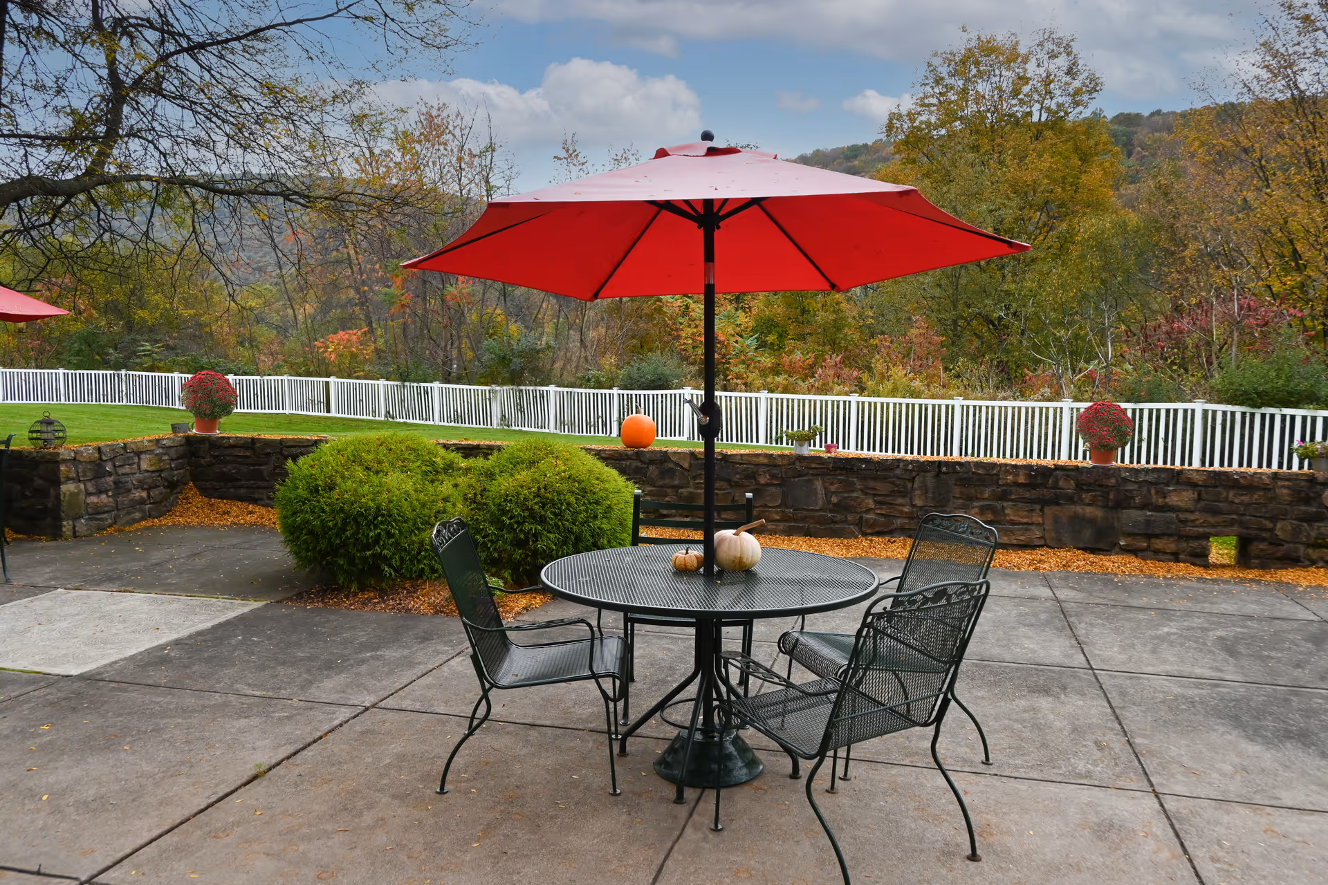 Outdoor patio area with a round metal table and four matching chairs under a large red umbrella. Three small pumpkins are placed on the table. The patio is surrounded by a low stone wall with potted plants and a white fence. Trees with autumn foliage and a cloudy sky are visible in the background.