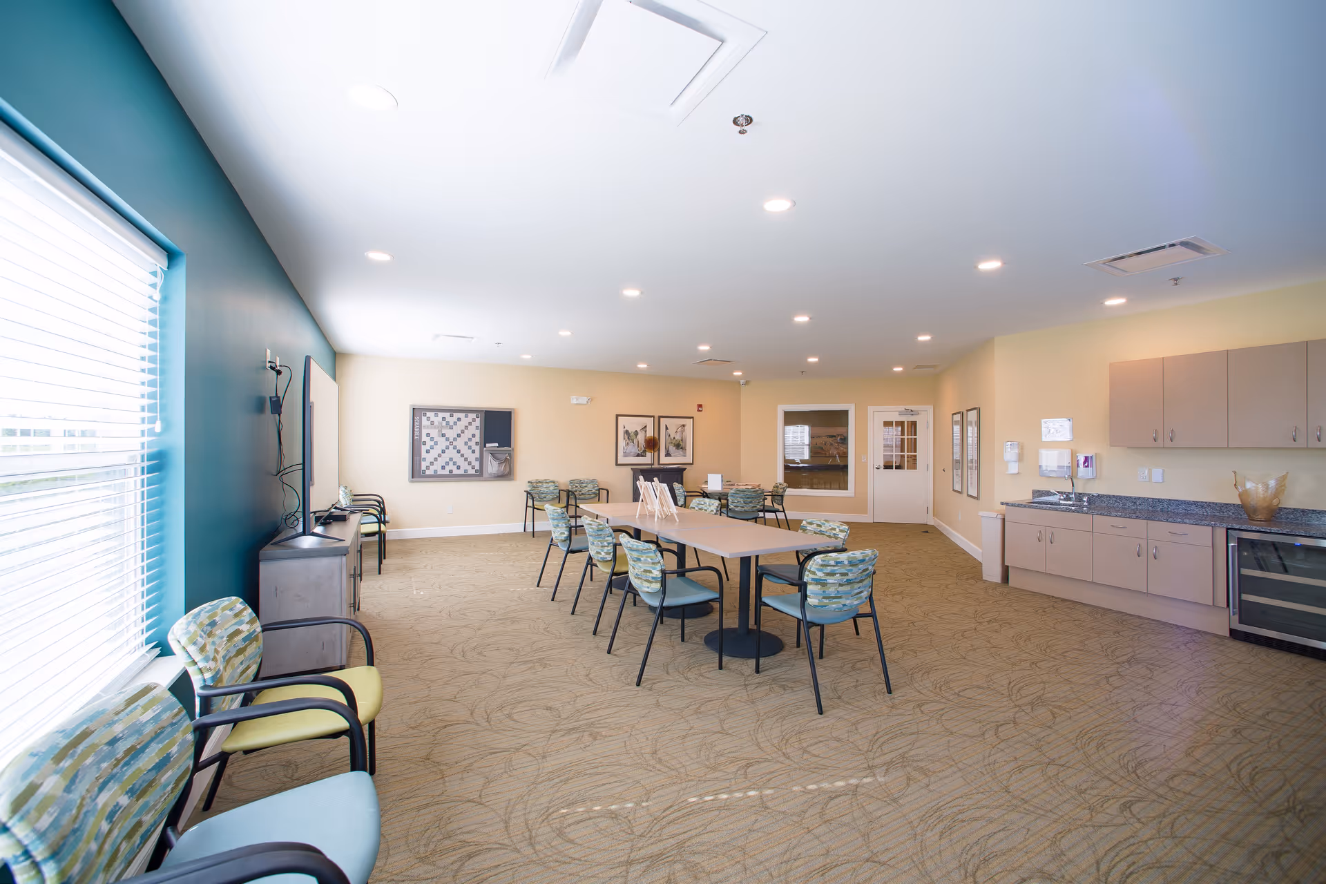 A spacious, well-lit common room with beige walls and carpeted floor. The room features a long table with several patterned chairs around it, additional chairs lined up against the walls, a large window with blinds, a mounted TV, and a game board on the wall. There is also a kitchenette area with cabinets, a countertop, and a wine cooler.
