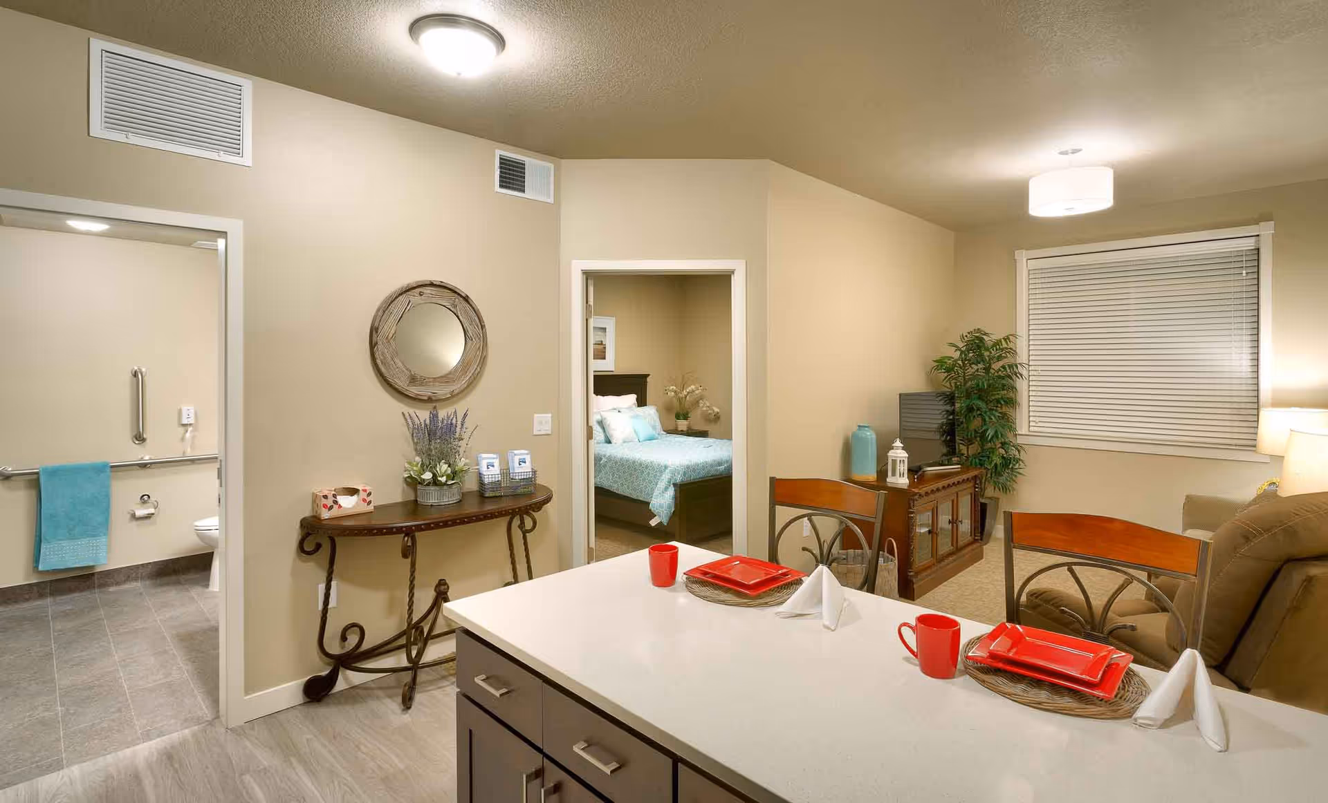 Open-plan senior apartment interior with a kitchen island set for two, a living area, and views into a bedroom and an accessible bathroom.