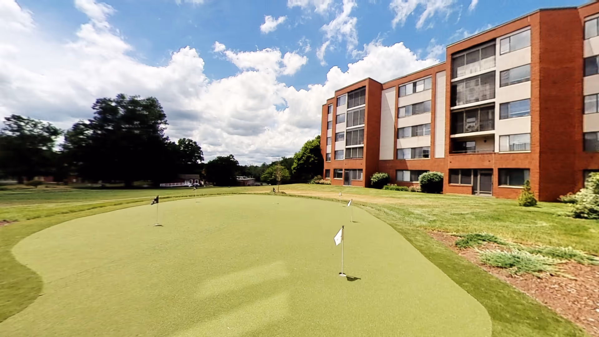A brick multi-story senior living building beside a putting green and lawn under a partly cloudy sky.