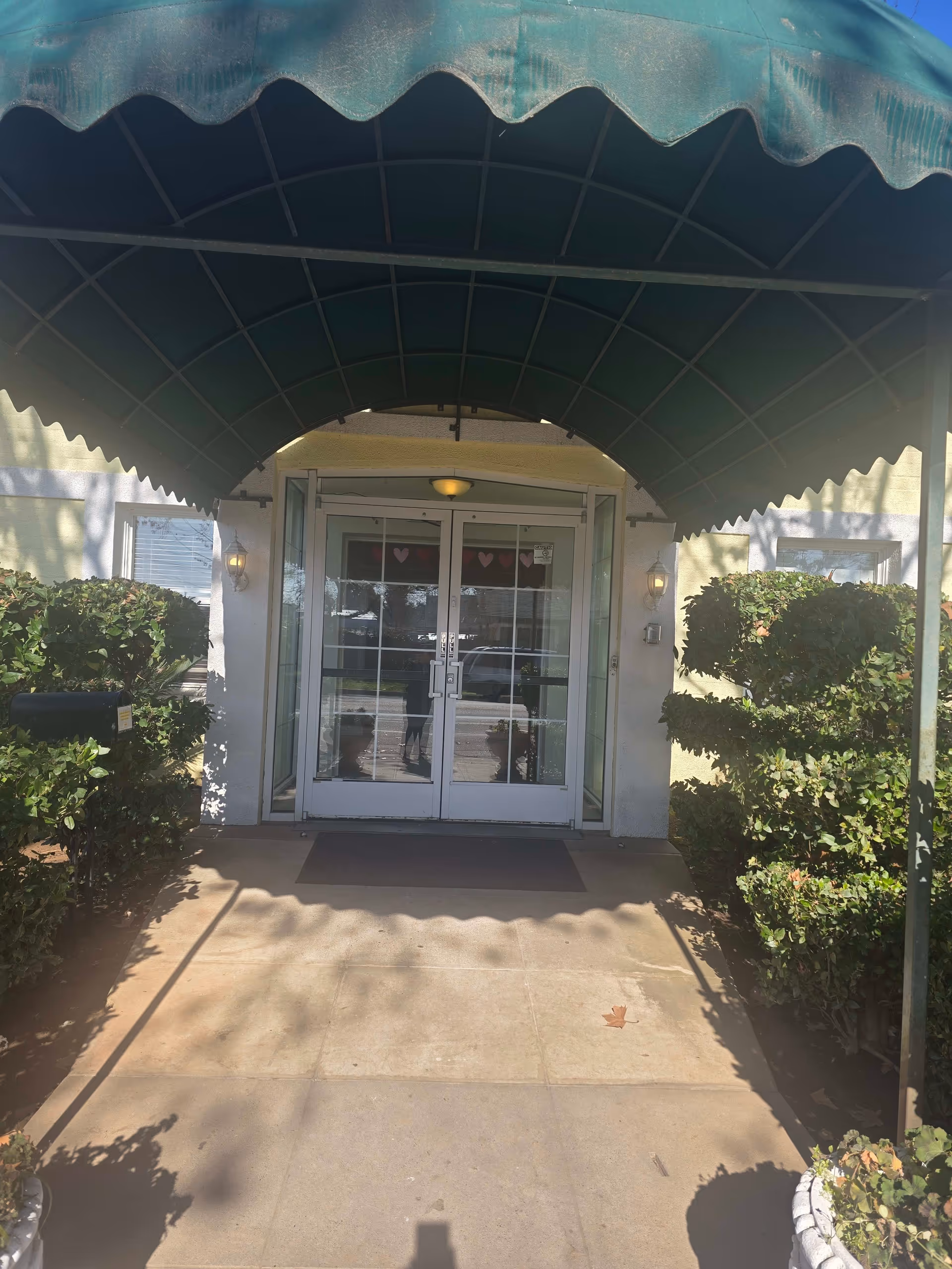 Entrance to a building with double glass doors under a green awning. There are trimmed bushes on either side of the walkway leading to the doors, and a mailbox is visible on the left side. The building exterior is light-colored with two wall-mounted lights flanking the doors.