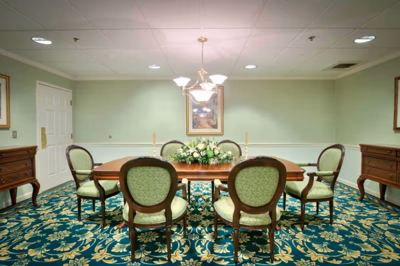 Formal dining room with a wooden table surrounded by six green upholstered chairs, a floral centerpiece, chandelier, and patterned carpet.