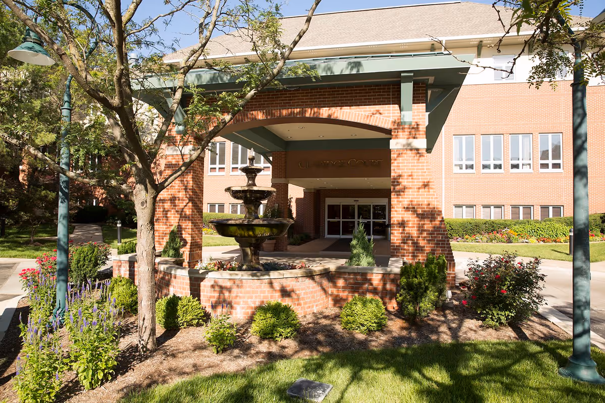 Entrance of Claridge Court facility with a brick exterior, a covered entryway, a tiered water fountain in front, and landscaped garden beds with shrubs and flowers. Trees and lamp posts line the walkway leading to the entrance.
