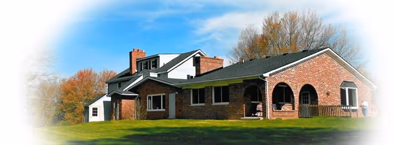 A large brick building with multiple sections and a dark roof, surrounded by green grass and trees with autumn foliage under a blue sky with some clouds.
