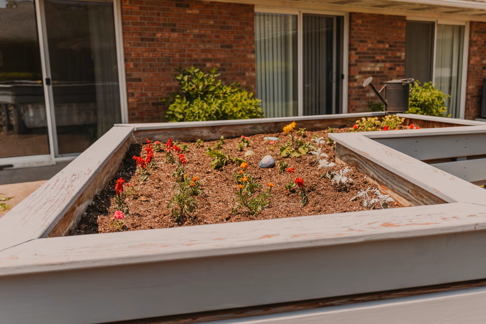 A raised garden bed with various small flowering plants and a watering can placed on the edge. The garden bed is situated outside in front of a brick building with sliding glass doors and windows.