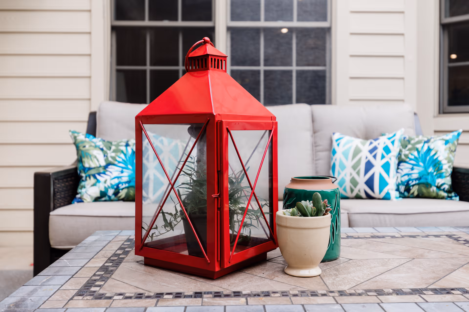 A red decorative lantern and potted succulents sit on a tiled patio table in front of a cushioned outdoor sofa with patterned pillows.